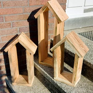 May include: Three house-shaped wooden frames constructed from light-colored natural wood planks. Each frame features a peaked roof design with vertical side walls and a flat base. The wood displays visible grain patterns and light brown tones.