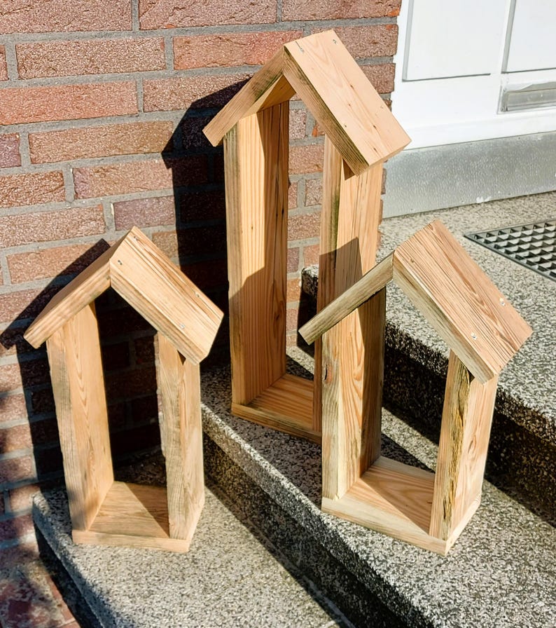 May include: Three house-shaped wooden frames constructed from light-colored natural wood planks. Each frame features a peaked roof design with vertical side walls and a flat base. The wood displays visible grain patterns and light brown tones.