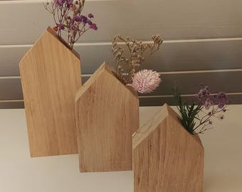 May include: Three light-colored wooden house-shaped vases of varying heights, each featuring a peaked roof design. Each wooden block contains a small opening at the top holding delicate dried flowers in shades of purple, pink, and beige.