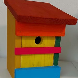 Könnte beinhalten: Ein bunt bemaltes Vogelhaus aus Holz mit rotem Dach, orangefarbenen Zierleisten und gelbem Korpus. Das Vogelhaus hat ein rundes Einflugloch und ist mit pinkfarbenen, grünen und blauen Details verziert. Ein dekorativer Artikel für den Garten.