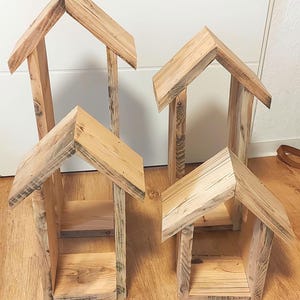 May include: Four house-shaped wooden structures of varying heights, each featuring a peaked roof and an open front design. The natural light-colored wood displays visible grain patterns and textures, with each piece constructed from vertical planks.