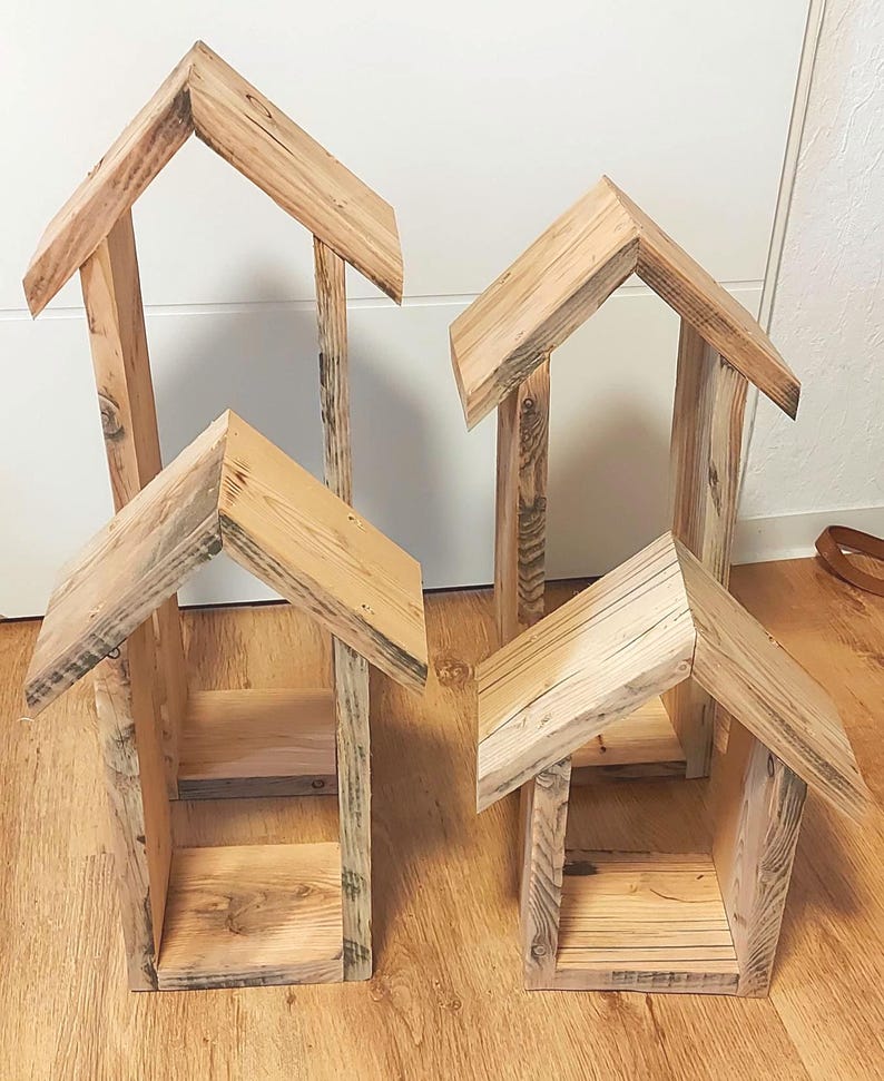 May include: Four house-shaped wooden structures of varying heights, each featuring a peaked roof and an open front design. The natural light-colored wood displays visible grain patterns and textures, with each piece constructed from vertical planks.