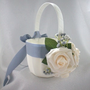 May include: A white flower girl basket with a blue satin ribbon and a white rose and blue flower embellishment.