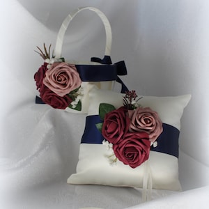 May include: A white satin pillow with a navy blue ribbon and a bouquet of burgundy and blush roses. The pillow is for a wedding ceremony.