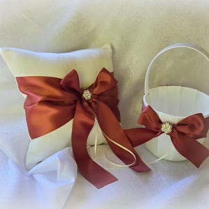 May include: A white ring bearer pillow and flower girl basket set. The pillow and basket are adorned with burgundy satin bows and sparkling rhinestone accents. The set is designed for a wedding.