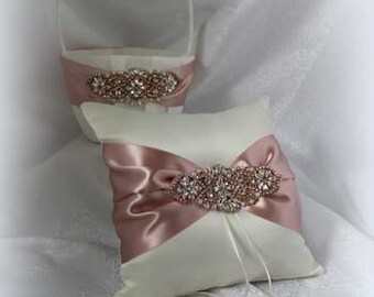 May include: A white satin ring pillow and matching flower girl basket. Both items feature a blush pink satin ribbon and a decorative rhinestone embellishment. The basket has a handle and the pillow has ribbons.