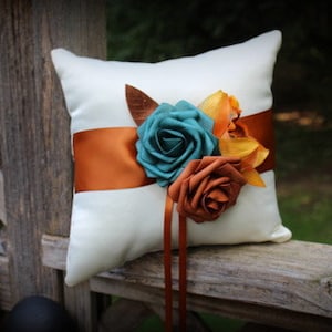 May include: A white satin ring pillow with a rust-colored satin ribbon and three fabric flowers. The flowers are teal, rust, and yellow.