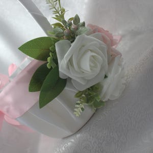 May include: A white satin flower girl basket with a pink satin bow and a bouquet of white and pink roses with green leaves.