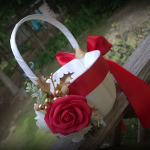 May include: White flower girl basket with a red satin ribbon and gold accents. The basket is decorated with a red rose, gold leaves, and white berries.