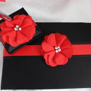 May include: Black guest book and pen holder set, adorned with red chiffon flower accents. The guest book features a red ribbon and a matching pen. Each flower is embellished with rhinestones and pearls.