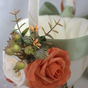 May include: A white flower girl basket with a green satin ribbon and a bouquet of orange and white flowers. The basket is decorated with green leaves and berries.