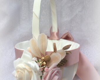 May include: A white and pink flower girl basket with a handle. The basket is decorated with cream and pink roses, leaves, and gold accents. A pink ribbon is tied around the basket and handle.