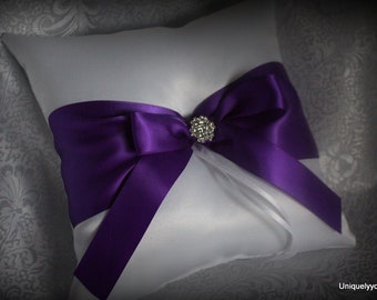 May include: White satin ring pillow with a large purple satin bow and a rhinestone embellishment in the center.