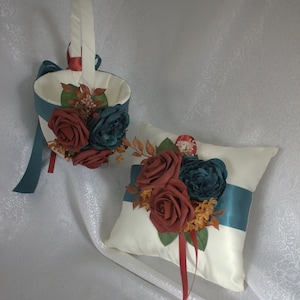 May include: A white satin pillow with teal and burgundy ribbon and a floral arrangement of burgundy and teal roses. The pillow is for a wedding ceremony.