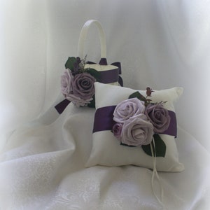 May include: A white satin ring pillow with a purple satin ribbon and three lavender roses. A white basket with a purple ribbon and lavender roses is in the background.