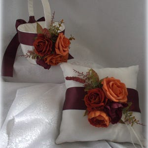 May include: A white satin pillow with a burgundy satin ribbon and a bouquet of orange and burgundy silk flowers. A white satin flower girl basket with a burgundy satin ribbon and a bouquet of orange and burgundy silk flowers.