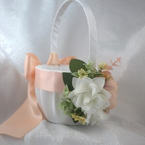 May include: A white satin flower girl basket with a peach satin ribbon and a bouquet of white and peach flowers.
