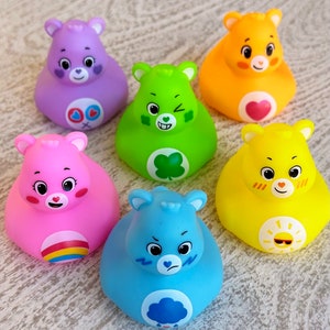 May include: Six colorful rubber duckies shaped like Care Bears. Each duck has a different color and a different Care Bear symbol on its chest. The ducks are arranged in a circle on a wooden surface.
