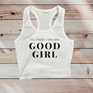 Pode incluir: Top branco com a frase "Yes, Daddy I am your GOOD GIRL" em preto. O top tem decote redondo e design de costas nadador. O texto é impresso em uma fonte sem serifa em negrito. O top é exibido em uma superfície de madeira.