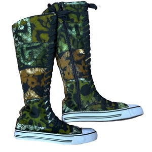 May include: Knee-high boots with a distinctive pattern. The boots have a green, brown, and gold design, black laces, and a zip. The soles are white with black stripes, and the toe caps are white.