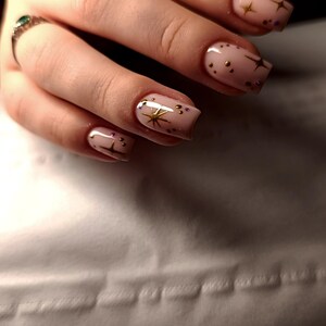 May include: Close-up of manicured nails with a light pink base. Each nail features gold star and dot designs. A silver ring with a green stone is visible on the ring finger.