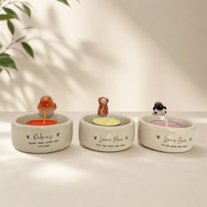 May include: Three cream-coloured ceramic candle holders, each with a small animal figurine and a lit tealight candle. The text on the holders reads: 'Robins', 'Love Moo', and 'Love Eve'.