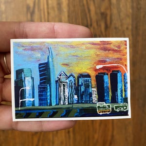 May include: A rectangular sticker featuring a painted city skyline at sunset with vibrant yellow, orange, and blue hues. The scene depicts dark blue skyscrapers against a bright sky, with a green highway sign in the foreground reading Central Phila.