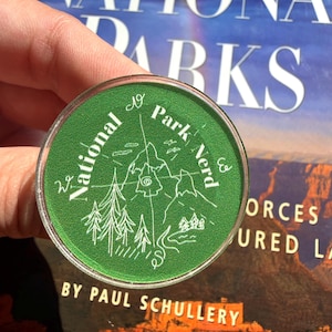 May include: A round, green button with the words "National Park Nerd" encircling a white illustration of mountains, trees, and a compass. The button is held in a hand, with the words "National Parks" visible in the background.