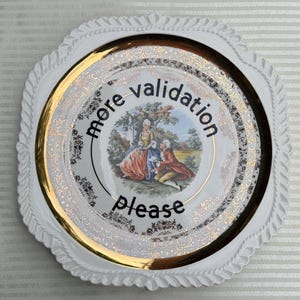 May include: A white decorative plate with a scalloped edge and a gold-trimmed border featuring an intricate floral pattern. The center displays a colorful illustration of two figures in period clothing with the black text more validation please.