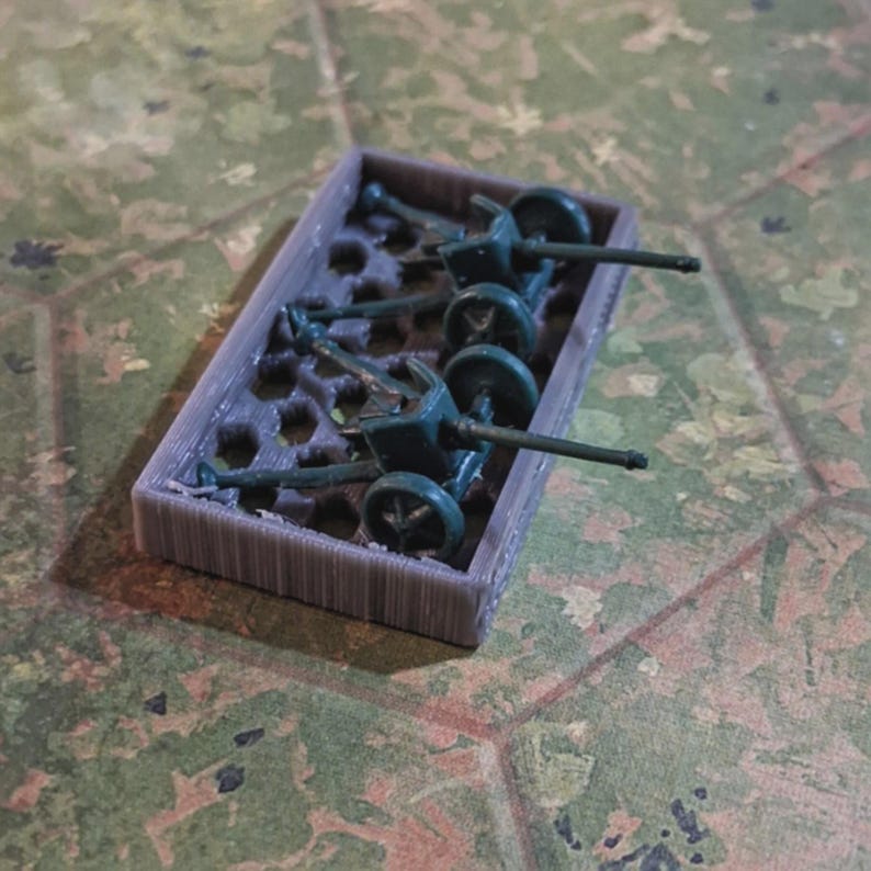 May include: A set of miniature green artillery pieces in a gray rectangular tray. The miniature cannons have wheels and long barrels. The tray has a honeycomb-like interior. The background is a camouflage pattern.