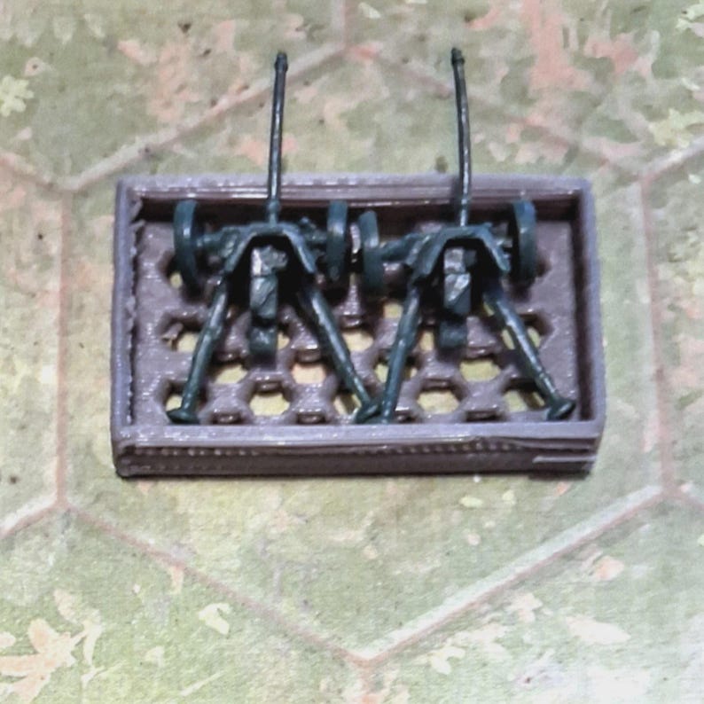 May include: Miniature model of two dark green artillery pieces mounted on a brown rectangular base. The artillery pieces have detailed gun barrels and support structures, set against a backdrop of a hexagonal pattern.