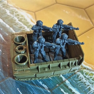 Könnte beinhalten: Ein miniaturisiertes olivgrünes Amphibienfahrzeug, das vier graue Soldatenfiguren trägt, die jeweils ein Gewehr halten. Das Fahrzeug hat drei kreisförmige Öffnungen an der Seite und befindet sich auf einem Spielbrett mit Wasser- und Sanddesign.