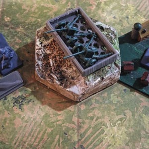 May include: A tabletop game scene with three hexagonal game board sections. One section features gray structures and blue figures. Another holds green plastic soldiers in a container. The third has wooden structures and figures on a green surface.