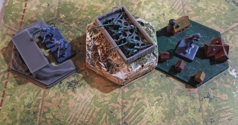 May include: A tabletop game scene with three hexagonal game board sections. One section features gray structures and blue figures. Another holds green plastic soldiers in a container. The third has wooden structures and figures on a green surface.