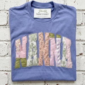 May include: A periwinkle blue t-shirt with the word "NANNA" appliqued across the front in patchwork fabric. The patchwork includes floral, paisley, and gingham patterns in pink, green, and white. A label at the neckline reads "Chapel's Closet".