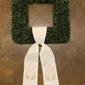 以下が含まれることがあります： 両側にピンクとグリーンで「EPI」のモノグラムが刺繍された白い布製のリースボウ。ボウは緑のツゲのリースに結ばれています。
