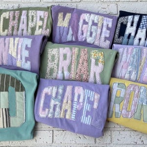 Peut inclure: Plusieurs sweatshirts de différentes couleurs, chacun avec un nom ou un numéro appliqué dans un style patchwork. Les noms et les chiffres sont dans une police universitaire, avec une variété de tissus à motifs utilisés pour les lettres et les chiffres.