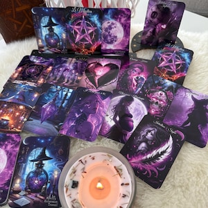 Peut inclure: Un jeu de cartes de tarot avec des illustrations mystiques dans des tons violets et bleus, représentant des lunes, des étoiles et des figures. Une bougie allumée dans un pot en verre est placée devant les cartes. Un livre relié en cuir est visible.
