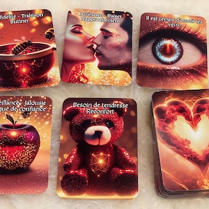 Op de afbeelding: Een pakje orakelkaarten met afbeeldingen en Franse tekst. De kaarten tonen illustraties van een bij in een kom, een kussend stel, een oog, een appel, een teddybeer en een hart. De kaarten hebben een warm, gloeiend kleurenschema.