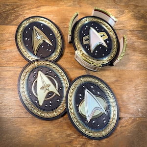 Peut inclure: Quatre dessous de verre ovales avec un design d'insigne Starfleet. Les dessous de verre ont un fond noir, une bordure dorée et un emblème doré et blanc. Fabriqués dans un matériau durable, ils protègent les surfaces des déversements.