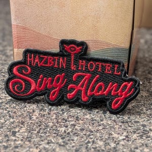 Peut inclure: Un écusson brodé noir avec le texte rouge Hazbin Hotel Sing Along et une petite icône de trident rouge centrée au-dessus du texte. L'écusson présente une bordure noire épaisse et une surface en tissu texturé avec des coutures rouges.