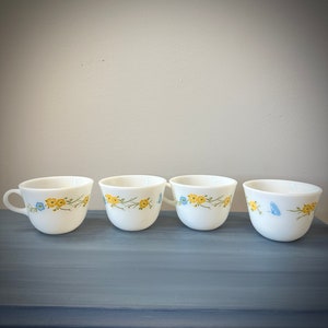 May include: Four white ceramic teacups with a floral design. The cups have a yellow and blue floral pattern with a green stem.