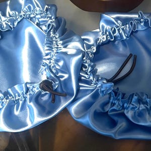 Op de afbeelding: Twee lichtblauwe satijnen mutsen met elastische trekkoorden. De mutsen hebben een gerimpelde rand en een glanzende, gladde textuur. De mutsen zijn ontworpen om het haar te beschermen en kapsels te behouden.