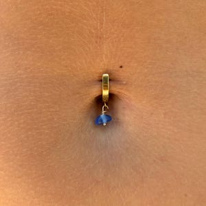 Puede incluir: Un piercing de ombligo dorado con una pequeña cuenta azul colgante. El anillo tiene un diseño de aro simple, y la cuenta es de color azul claro. La piel alrededor del ombligo es de color marrón claro.