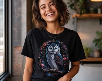 May include: Black t-shirt featuring a detailed illustration of an owl perched on a branch, surrounded by a floral wreath. The owl has white, brown, and blue feathers. The shirt is a classic crew neck style.