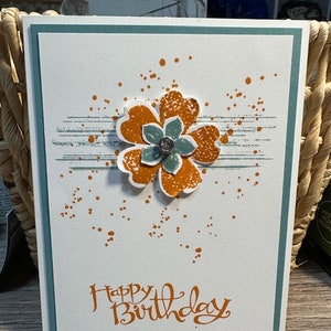 May include: A handmade birthday card with a white background, teal border, and a layered flower. The flower has orange petals and a teal centre with a gem. The words "Happy Birthday" are written in orange script.