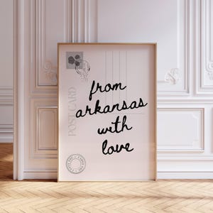 Może przedstawiać: Oprawiony nadruk pocztówki z napisem "from arkansas with love" czarnym pismem. Pocztówka ma wzór znaczka pocztowego w lewym górnym rogu i okrągłą pieczęć na dole. Rama jest w kolorze jasnobrązowym.