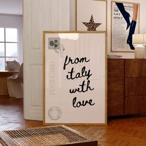 Op de afbeelding: Een ingelijste print met de tekst "from italy with love" in cursief schrift, ontworpen als een ansichtkaart. De print bevat een postzegel en een rond zegel. Andere ingelijste kunstwerken zijn zichtbaar, waaronder een ster met luipaardprint.