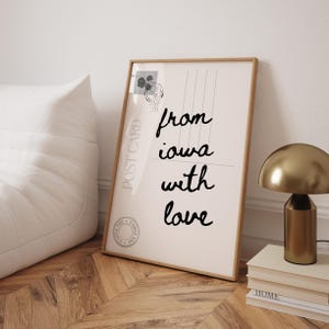 May include: A framed print with the words "from Iowa with love" in black script. The print has a vintage postcard design with a stamp and a "Post Card" label. A gold-colored lamp and books are on a wooden surface.