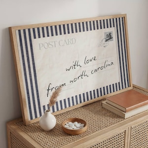 May include: A framed rectangular print featuring a vintage postcard design with dark blue and white vertical stripes on the edges. The center displays the text POST CARD, a small stamp graphic, and the handwritten script with love from north carolina.